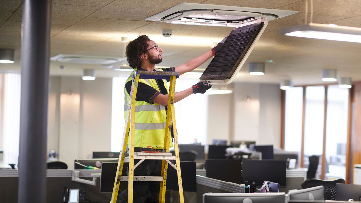 Technicien sur escabeau remettant en place la grille d'une climatisation de bureau